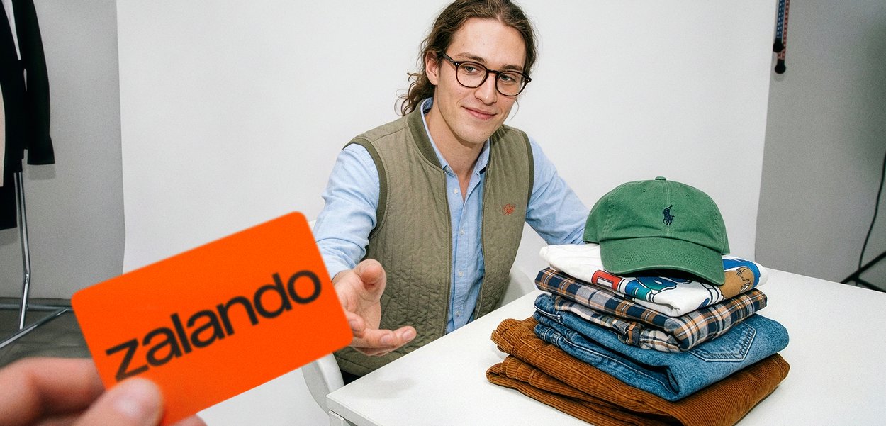 Hombre con gafas y chaleco sentado en una mesa con ropa doblada y gorra verde, alcanzando una tarjeta regalo Zalando de color naranja brillante.
