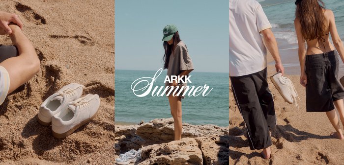 Hvide sneakers på sand ved siden af en persons fod og en kvinde i en casual t-shirt og kasket, der står på klipper ved havet under klar himmel.