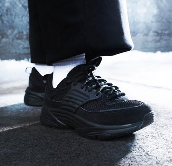 Chaussures de sport noires au design texturé portées avec des chaussettes blanches et un pantalon noir sur un sol en béton sombre.