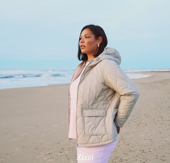 Kvinde iført en beige quiltet hættejakke og lyserøde bukser, stående på en sandstrand og kiggende mod havet.