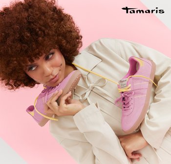 Frau mit lockigem Haar trägt eine beige Jacke und hält ein Paar pinke Sneakers mit gelben Schnürsenkeln, die sie gegen einen rosa-weißen Hintergrund hält und daran riecht.