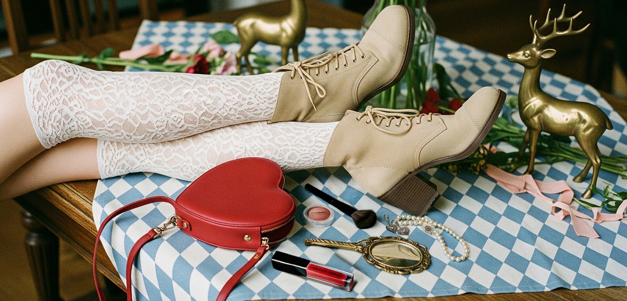 Beige lace-up ankle boots with a chunky heel, white lace thigh-high stockings, a red heart-shaped handbag, and assorted makeup items on a patterned tablecloth.
