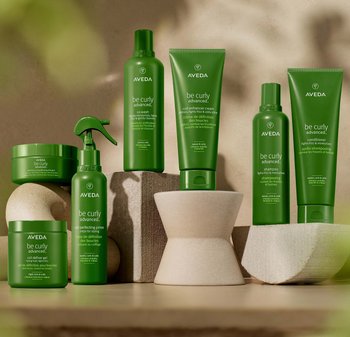 Aveda Be Curly Advanced haarproducten tentoongesteld. Flesjes en potten in verschillende schakeringen groen, met een matte textuur, en met cream- en sprayapplicators.