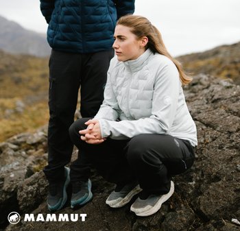 Hellgraue Daunenjacke mit hohem Kragen, schwarze Hose und graue Schuhe. Außenbereich mit felsigem Gelände und verschwommenem Hintergrund.