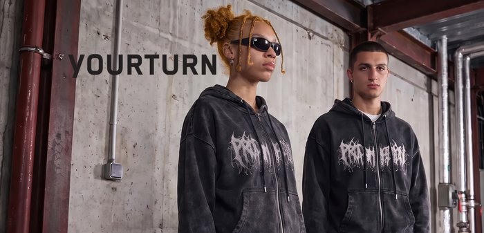 Two young people wearing matching black graphic hoodies stand side by side in an industrial setting with exposed pipes and a concrete wall.