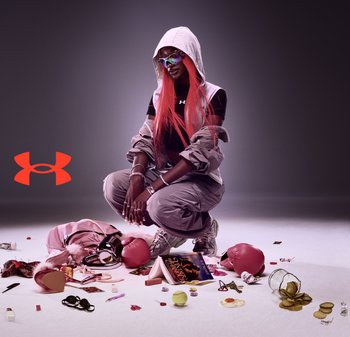 Persoon met lang roze haar, zonder mouwen jas met capuchon en zonnebril hurkt te midden van verspreide voorwerpen waaronder bokshandschoenen, boek, tennisbal, omgevallen augurken en make-up.