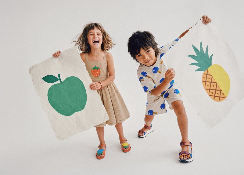 Twee kinderen glimlachen en houden grote papieren borden vast met een illustratie van een groene appel en een ananas op een witte achtergrond.