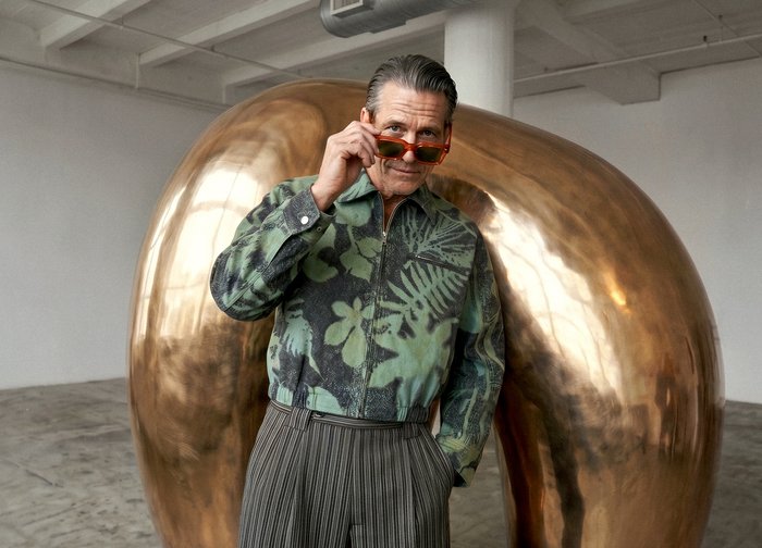 Homme en veste verte à motifs et pantalon rayé ajustant des lunettes de soleil orange devant une grande sculpture abstraite en bronze brillant dans un espace de galerie.