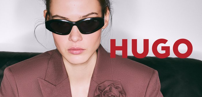 Schwarze Sonnenbrille mit einem schlanken, länglichen Design. Das moderne Gestell hat eine glänzende Oberfläche und passt gut zu einem braunen Blazer mit floralen Details.