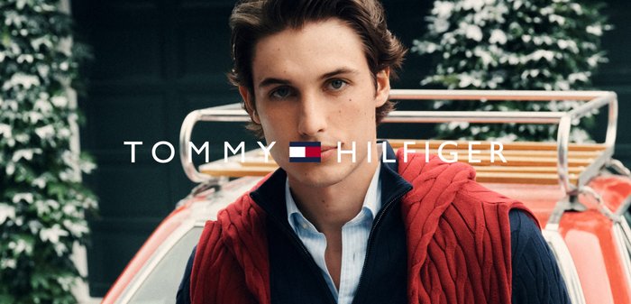 Männliches Model trägt einen roten Kabelstrickpullover über einem blauen Hemd. Der Hintergrund zeigt Grünflächen und ein Oldtimerauto. Logo unten angezeigt.