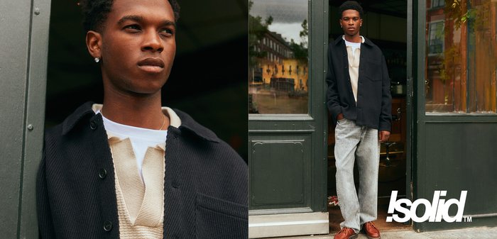 Marineblaues Hemd über einem cremefarbenen Strickpullover, hellgraue Jeans und braune Schuhe. Das Outfit wird durch einen schlichten Ohrring ergänzt.