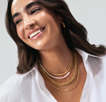 Colliers superposés en or et perles, présentant des designs de chaînes variés, sur une chemise blanche. Textures lisses et éclat subtil.