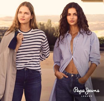 Two women stand outdoors wearing casual striped tops and jeans, one with a jacket over her shoulder, the other with hands in pockets.