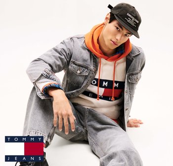 Denim jacket and jeans in light wash, layered over an orange hoodie and graphic sweater with red, blue, and white stripes. Black cap with logo.