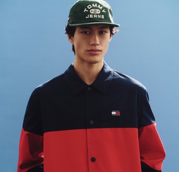 Joven con una chaqueta Tommy Jeans negra y roja y una gorra Tommy Jeans verde oliva sobre un fondo azul liso.