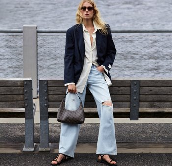 Marineblazer, witte blouse met knopen, lichtblauwe versleten wijde jeans, zwarte platte sandalen en een bruine handtas met patroon. Buitenomgeving.