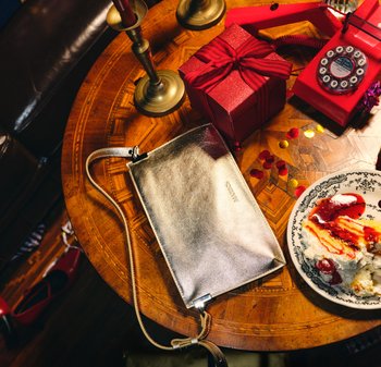 Metalicznie srebrna kopertówka z beżowym paskiem, o gładkiej fakturze. Leży na drewnianym stole obok czerwonego pudełka i zabytkowego telefonu.