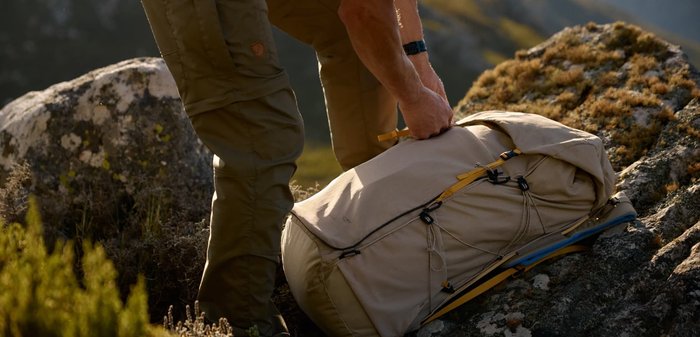 Person tightening straps on a beige hiking backpack resting on rocky terrain in a mountainous outdoor setting.