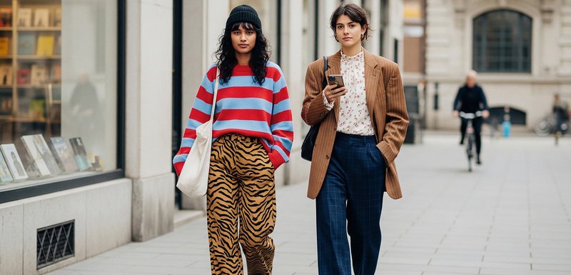 Deux femmes marchent dans une rue de la ville. L'une porte un pull rayé rouge et bleu et un pantalon à imprimé tigre. L'autre porte un blazer brun à rayures et un pantalon à carreaux.