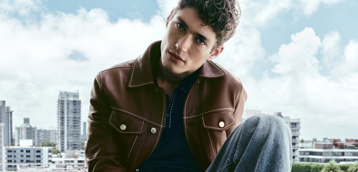 Ung mann i brun skinnjakke og jeans som sitter med byens skyline og skyet himmel i bakgrunnen.