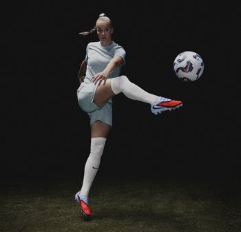 Atleta in maglia azzurro chiaro e pantaloncini bianchi, con calze bianche e scarpini rossi/blu, che calcia un pallone da calcio a motivi contro uno sfondo scuro.