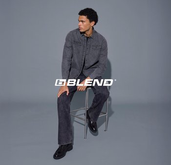 Graue Jeansjacke mit zwei Brusttaschen, kombiniert mit dunklen Jeans und schwarzen Schuhen, sitzend auf einem Metallhocker vor einem grauen Hintergrund.