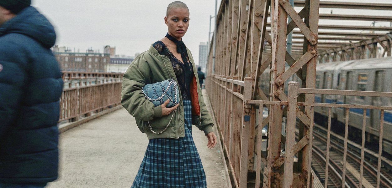 Mlada ženska s kratkimi lasmi, oblečena v zeleno jakno in karo krilo, drži modro prešito torbo na mestnem mostu blizu mimo vozečega vlaka.