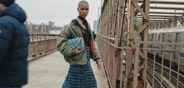 Junge Frau mit kurzen Haaren, die eine grüne Jacke und einen karierten Rock trägt, hält eine blaue gesteppte Tasche auf einer Stadtbrücke nahe eines vorbeifahrenden Zugs.