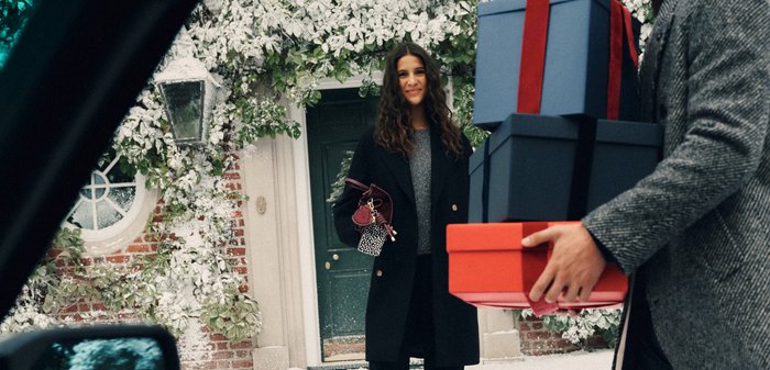 En person håller röda och blå presentlådor med band. Bakgrunden har en tegelvägg, en grön dörr och snötäckta växter.