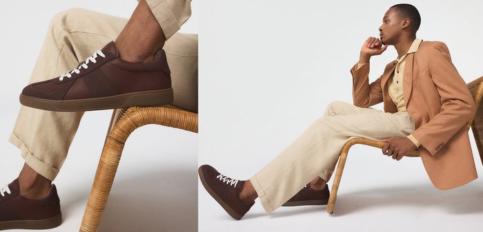 Mężczyzna w beżowych spodniach i brązowej marynarce balansuje na plecionym krześle, nosząc ciemnobrązowe trampki z białymi sznurówkami na jednolitym tle.