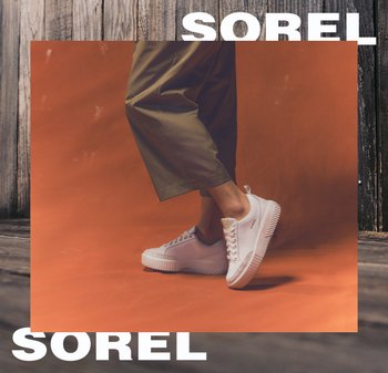 Person trägt weiße Sorel-Turnschuhe mit dicken Sohlen und olivgrüne Hosen, steht auf einer orangefarbenen Oberfläche vor einem hölzernen Hintergrund.