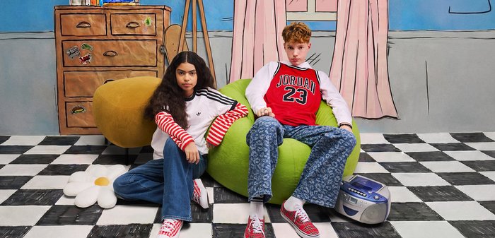 Deux adolescents, l'un assis sur un pouf vert et l'autre par terre, dans une pièce avec un sol en damier et une commode en bois.