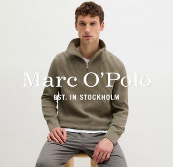 Mężczyzna w oliwkowo-zielonym swetrze zapinanym na zamek z prążkowanej dzianiny oraz szarych spodniach, siedzący na drewnianym stołku na tle jednolitego jasnego tła.