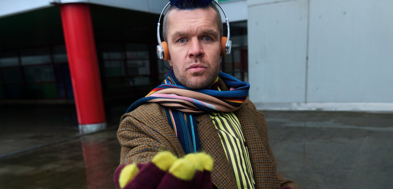 Mies, jolla on päässään kuulokkeet ja monivärinen raidallinen huivi, ruskea ruudullinen takki ja sormettomat hansikkaat, joiden kärjet ovat keltaiset, ojentaa kättään.