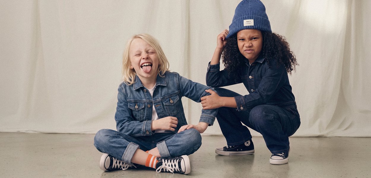 Due bambini vestiti in denim giocano in modo giocoso; uno è seduto con la lingua fuori, l’altro è accovacciato con il sopracciglio aggrottato e un berretto blu.