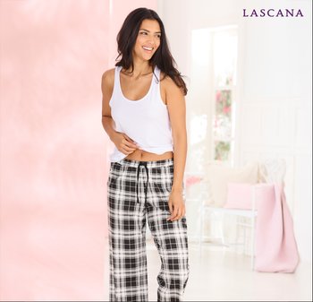 Glückliche vrouw in een witte tanktop en zwart-wit geblokte pyjamabroek, staand in een helder roze en witte kamer met een gezellige stoel.