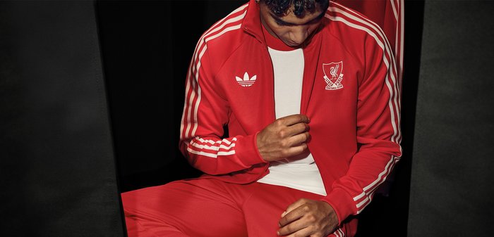Άνδρας που φοράει κόκκινη φόρμα Adidas με το έμβλημα της Liverpool FC, καθισμένος και κοιτάζει προς τα κάτω ενώ ρυθμίζει το φερμουάρ του μπουφάν.