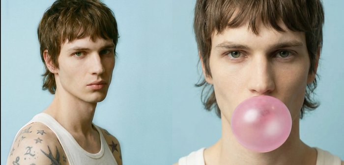 Jeune homme tatoué portant un débardeur blanc, soufflant une bulle de chewing-gum rose sur un fond bleu clair.