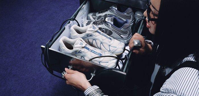 Person with glasses opening a metal drawer containing two pairs of athletic shoes and a microphone on a dark carpeted floor.