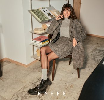 Geruit bruin blazer en rok in een wolmix; grijze coltrui; zwarte loafers; zittend op een houten stoel met een moderne boekenkast op de achtergrond.
