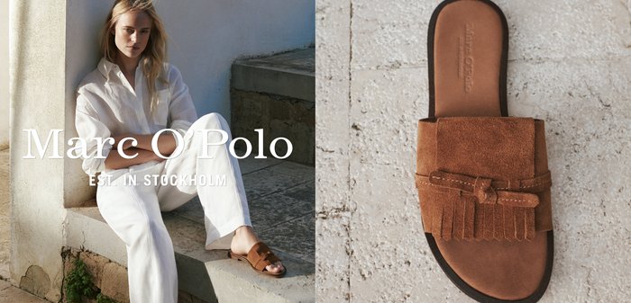 Donna con abito bianco seduta all'aperto che indossa sandali scivolati in camoscio marrone; primo piano di un sandalo scivolato in camoscio marrone con dettaglio di frange.