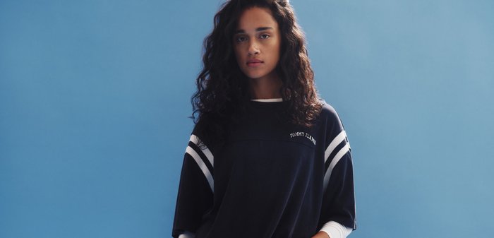 Mujer joven con cabello rizado que lleva una sudadera negra de Tommy Jeans, de pie contra un fondo azul liso.