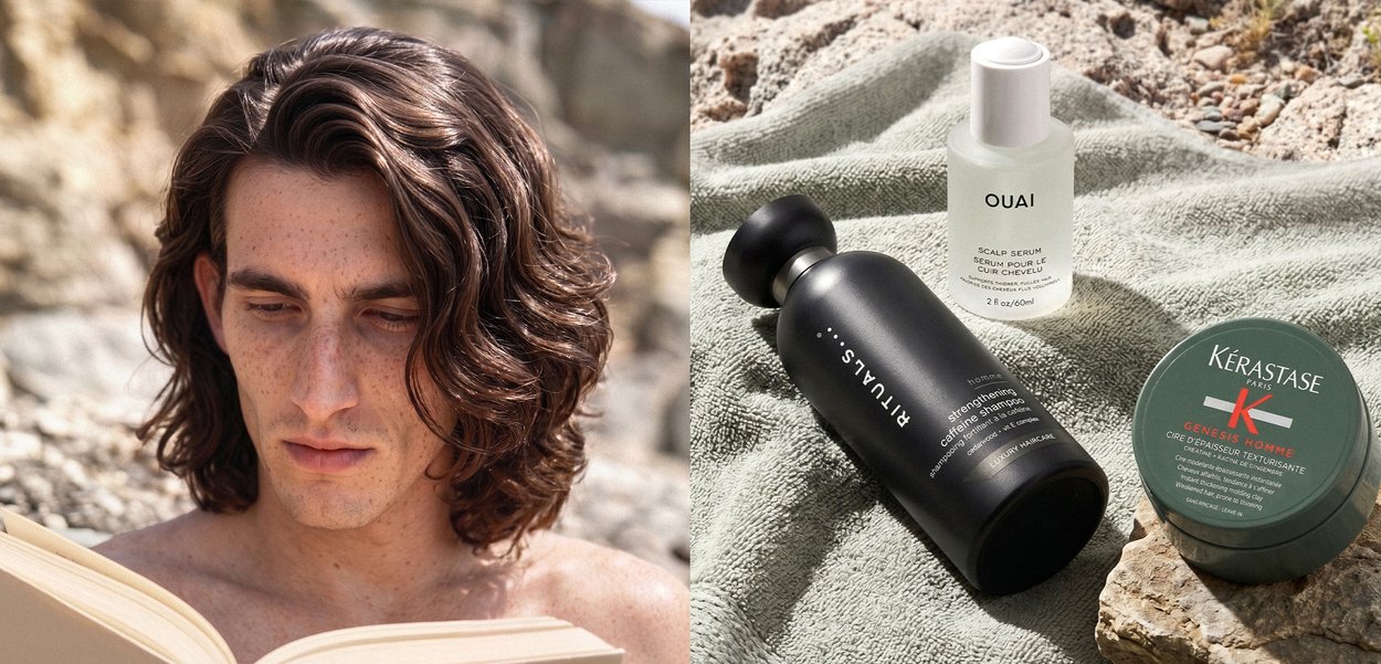 Mann mit welligem Haar liest draußen ein Buch neben einem Serum für die Kopfhaut, stärkendem Haarergänzungsmittel und texturierender Creme auf einem Handtuch und einer Steinoberfläche.