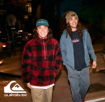 Deux jeunes hommes à l'extérieur la nuit. L'un porte un pull en polaire écossais rouge et noir ; l'autre a une veste à capuche bleue et un bonnet beige.