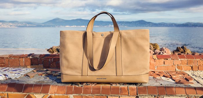 Bolso tote grande de color beige con asas dobles apoyado sobre un borde de ladrillo junto a un mar tranquilo bajo un cielo parcialmente nublado.