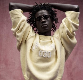 Persona che indossa occhiali da sole e un maglione giallo con scritte bianche, posa con le mani dietro la testa su uno sfondo rosa tenue.