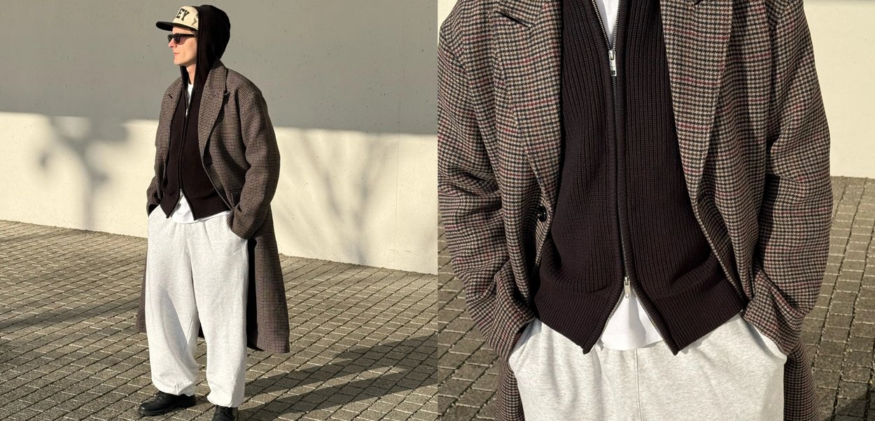 Mann iført rutete frakk, svart genser med glidelås, hvit skjorte, grå joggebukser, svarte sko, solbriller og svart caps som står på flislagt fortau.