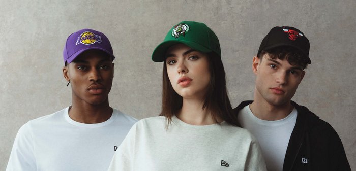 Tre giovani adulti che indossano cappellini sportivi casual con i loghi delle squadre NBA—Lakers, Celtics e Bulls—posano davanti a uno sfondo neutro.
