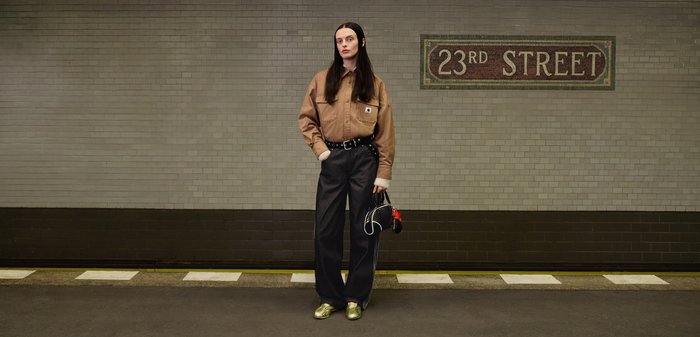 Personne aux longs cheveux foncés, vêtue d'une chemise marron, d'un pantalon noir et de chaussures dorées, tenant un petit sac noir sur un quai de métro sous le panneau 23rd Street.