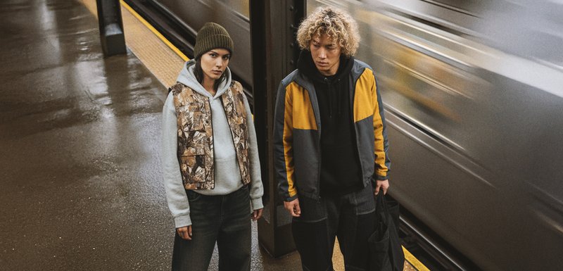 Deux personnes se tiennent à une station de métro. L'une porte un gilet camouflage par-dessus un sweat à capuche gris, tandis que l'autre arbore une veste en blocs de couleurs grise et jaune.